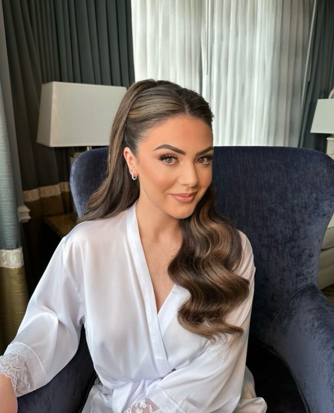 Bridal hair styling — sleek half-up with soft waves on a blue velvet chair.
